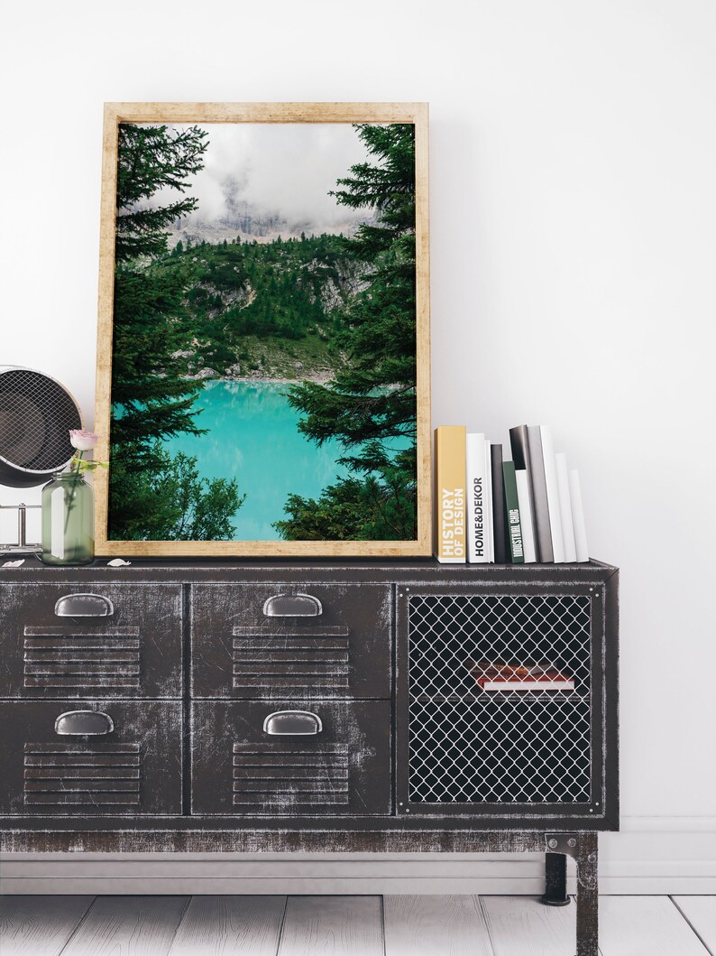 Op de afbeelding: Een ingelijste landschapsfoto van een meer omringd door bomen en bergen. Het meer is een levendige turquoise blauw en de bergen zijn bedekt met wolken. De foto hangt aan een muur boven een zwarte metalen kast met laden en een draadgaasdeur.