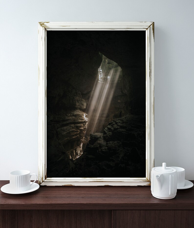 Op de afbeelding: Een ingelijste prent van een grotinterieur met zonlicht dat door een opening schijnt. De afbeelding zit in een wit, verweerde lijst en een witte kop en theepot staan op een donker houten oppervlak.