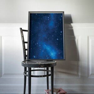 Pu&ograve; includere: Una stampa incorniciata nera di un cielo notturno blu scuro con una galassia bianca e stelle. La stampa &egrave; appoggiata su una sedia di legno nera con una pila di libri sul pavimento.