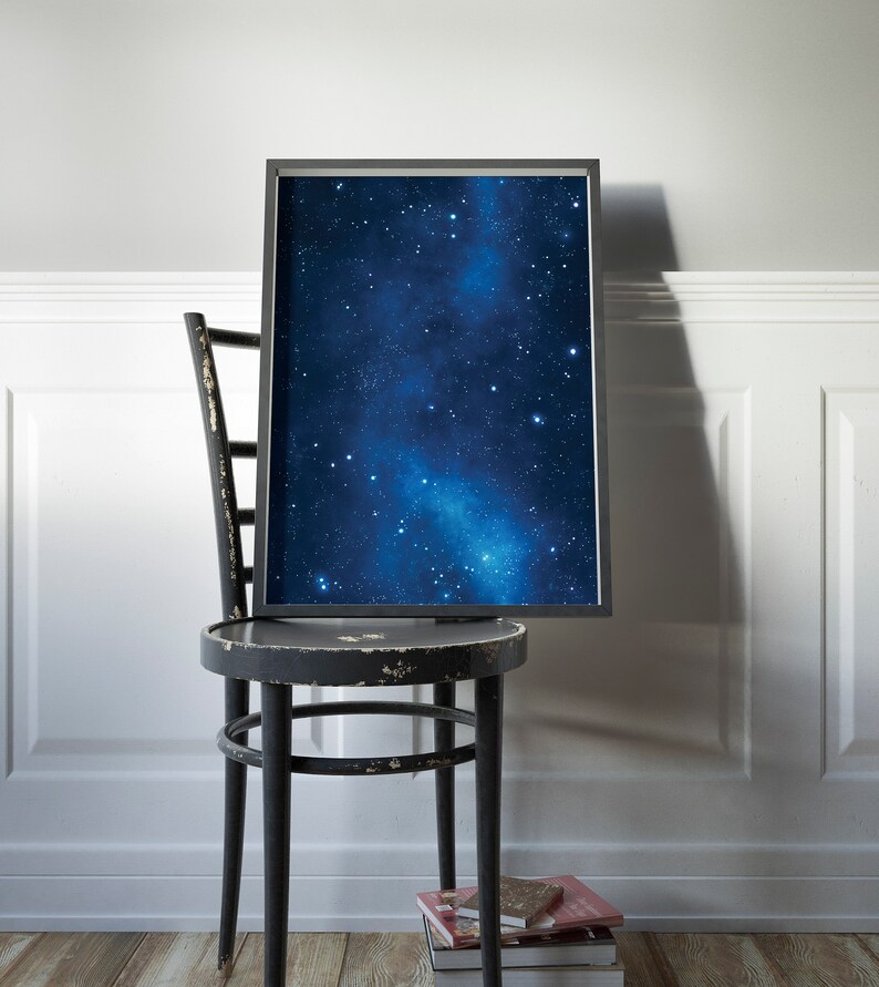 Pu&ograve; includere: Una stampa incorniciata nera di un cielo notturno blu scuro con una galassia bianca e stelle. La stampa &egrave; appoggiata su una sedia di legno nera con una pila di libri sul pavimento.