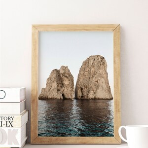 May include: A framed photograph of two large rock formations rising out of the ocean. The water is a deep blue and the sky is a light blue. The rocks are a light brown color.