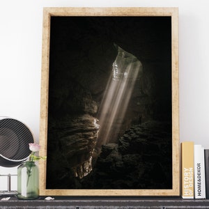 Op de afbeelding: Een ingelijste print toont een grotinterieur met zonlicht dat door een opening schijnt. De afbeelding toont een donkere, rotsachtige omgeving met een persoon in de verte. De lijst is licht houtkleurig en de print wordt tentoongesteld op een donkere metalen kast.