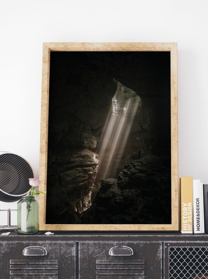 Op de afbeelding: Een ingelijste print toont een grotinterieur met zonlicht dat door een opening schijnt. De afbeelding toont een donkere, rotsachtige omgeving met een persoon in de verte. De lijst is licht houtkleurig en de print wordt tentoongesteld op een donkere metalen kast.