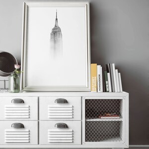 Peut inclure: Un meuble de rangement blanc en m&eacute;tal de style casier avec quatre tiroirs et une &eacute;tag&egrave;re en grillage m&eacute;tallique. Le meuble est dot&eacute; d'un cadre en m&eacute;tal noir et repose sur un sol en bois fonc&eacute;. Un imprim&eacute; noir et blanc encadr&eacute; de l'Empire State Building est accroch&eacute; au mur au-dessus du meuble. Une pile de livres est pos&eacute;e sur le meuble, dont un intitul&eacute; "Histoire du design".