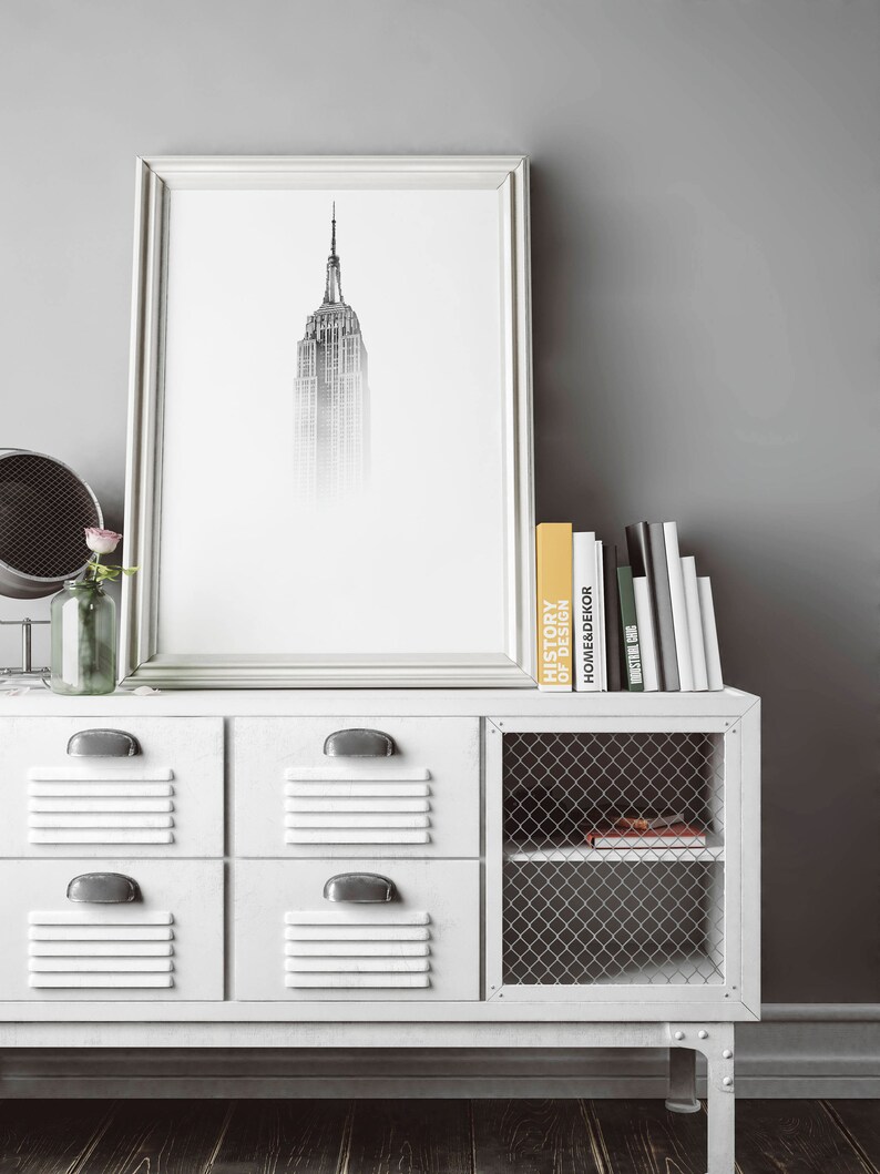 Peut inclure: Un meuble de rangement blanc en m&eacute;tal de style casier avec quatre tiroirs et une &eacute;tag&egrave;re en grillage m&eacute;tallique. Le meuble est dot&eacute; d'un cadre en m&eacute;tal noir et repose sur un sol en bois fonc&eacute;. Un imprim&eacute; noir et blanc encadr&eacute; de l'Empire State Building est accroch&eacute; au mur au-dessus du meuble. Une pile de livres est pos&eacute;e sur le meuble, dont un intitul&eacute; "Histoire du design".