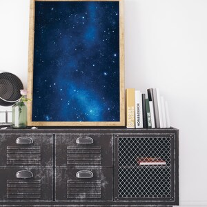 Pu&ograve; includere: Una stampa incorniciata di un cielo notturno blu scuro con una galassia bianca e stelle. La stampa &egrave; su un mobile in metallo nero con quattro cassetti e una porta in rete metallica. Il mobile &egrave; su un pavimento in legno bianco. Ci sono libri sopra il mobile, tra cui "History of Design" e "Home Decor".
