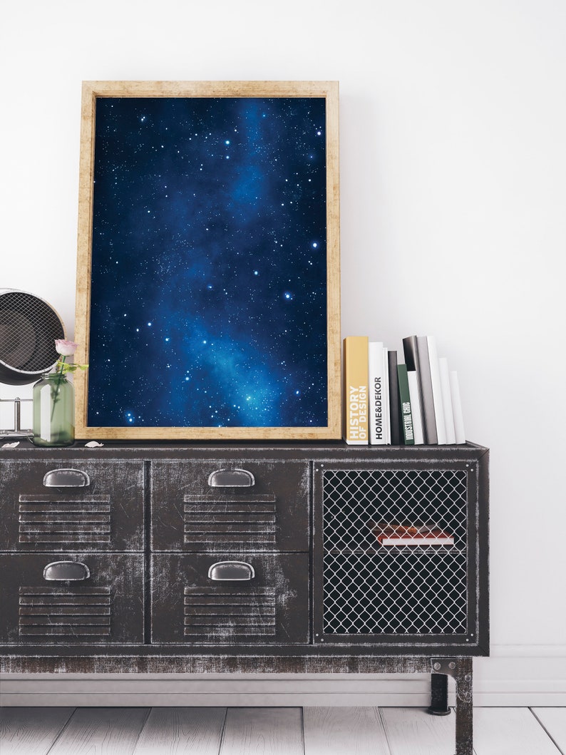 Pu&ograve; includere: Una stampa incorniciata di un cielo notturno blu scuro con una galassia bianca e stelle. La stampa &egrave; su un mobile in metallo nero con quattro cassetti e una porta in rete metallica. Il mobile &egrave; su un pavimento in legno bianco. Ci sono libri sopra il mobile, tra cui "History of Design" e "Home Decor".