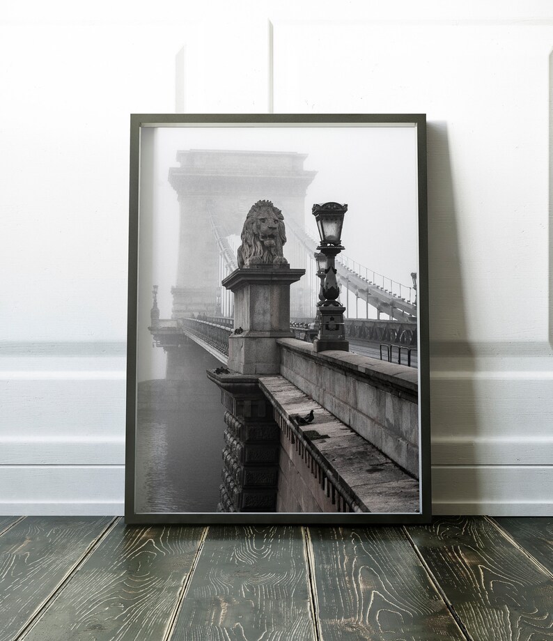 May include: A black and white photograph of a bridge with a lion statue on top of a pillar. The bridge is covered in fog and there is a street lamp on the side of the bridge.