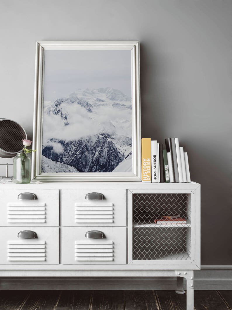 Peut inclure: Un meuble blanc avec quatre tiroirs et une &eacute;tag&egrave;re en fil m&eacute;tallique. Le meuble a un cadre blanc et des poign&eacute;es argent&eacute;es. Une photographie encadr&eacute;e d'une cha&icirc;ne de montagnes enneig&eacute;e est accroch&eacute;e au-dessus du meuble. Une pile de livres est pos&eacute;e sur le dessus du meuble, dont un avec le titre "Histoire du design".