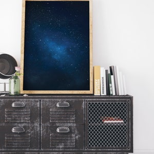 K&ouml;nnte beinhalten: Ein gerahmter Druck eines dunkelblauen Nachthimmels mit einer Galaxie von Sternen. Der Druck h&auml;ngt an einer wei&szlig;en Wand &uuml;ber einem schwarzen Metallschrank mit Schubladen und einer Gittert&uuml;r. Mehrere B&uuml;cher sind auf dem Schrank gestapelt.