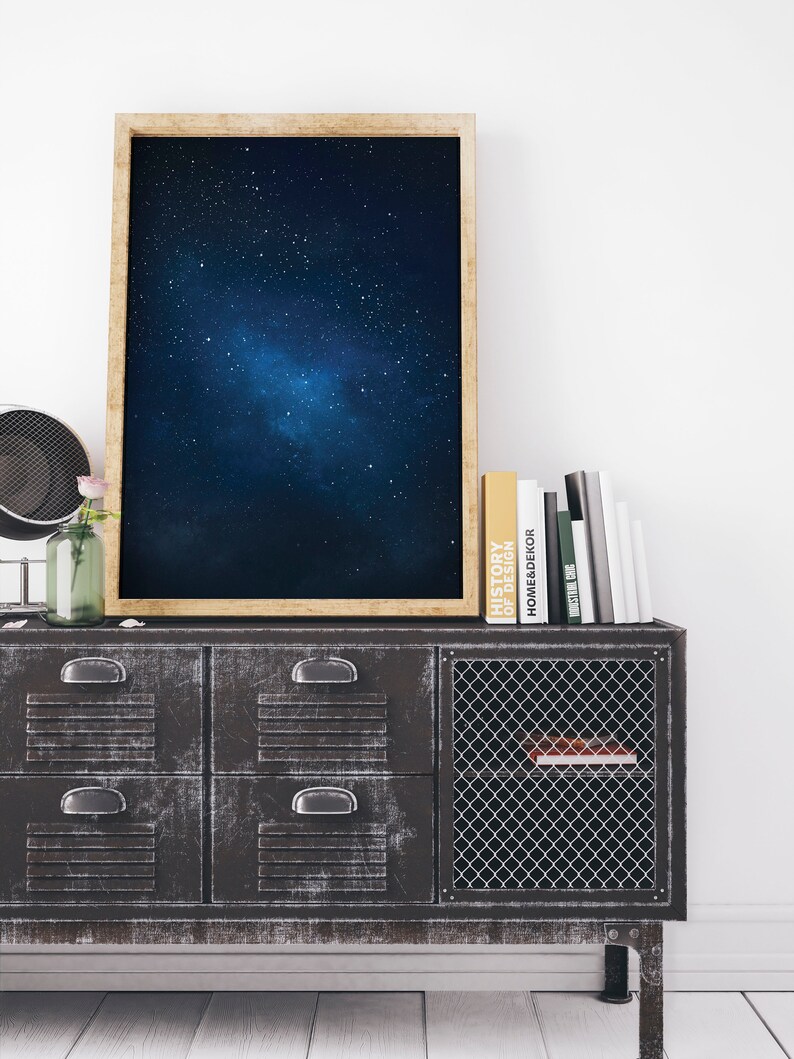 K&ouml;nnte beinhalten: Ein gerahmter Druck eines dunkelblauen Nachthimmels mit einer Galaxie von Sternen. Der Druck h&auml;ngt an einer wei&szlig;en Wand &uuml;ber einem schwarzen Metallschrank mit Schubladen und einer Gittert&uuml;r. Mehrere B&uuml;cher sind auf dem Schrank gestapelt.