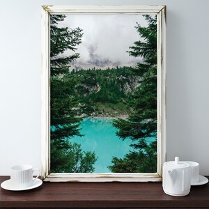 Op de afbeelding: Een ingelijste foto van een bergmeer omringd door bomen. Het meer is een levendig turquoise blauw en de bergen zijn bedekt met weelderige groene bomen. De foto is ingelijst in een wit verweerd frame.