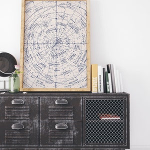 Puede incluir: Una impresi&oacute;n enmarcada de estilo vintage de una carta de estrellas con constelaciones y sus nombres en tinta azul sobre un fondo crema. La impresi&oacute;n est&aacute; colgada sobre un armario de metal negro con cuatro cajones y una puerta de malla de alambre. El armario est&aacute; sentado en un suelo de madera blanca.