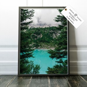 Op de afbeelding: Een ingelijste foto van een bergmeer omringd door weelderige groene bomen. Het meer is een levendig turquoise blauw en de bergen zijn bedekt met mist. De afbeelding is ingelijst in een zwart frame.