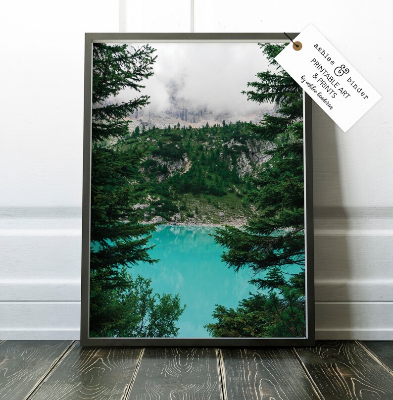 Op de afbeelding: Een ingelijste foto van een bergmeer omringd door weelderige groene bomen. Het meer is een levendig turquoise blauw en de bergen zijn bedekt met mist. De afbeelding is ingelijst in een zwart frame.