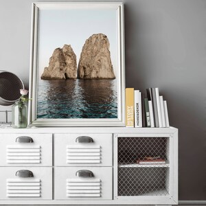 May include: A framed photograph of two large rock formations rising out of the ocean. The water is a deep blue and the sky is a light blue. The photo is hanging on a white wall above a white dresser with a wire mesh shelf.