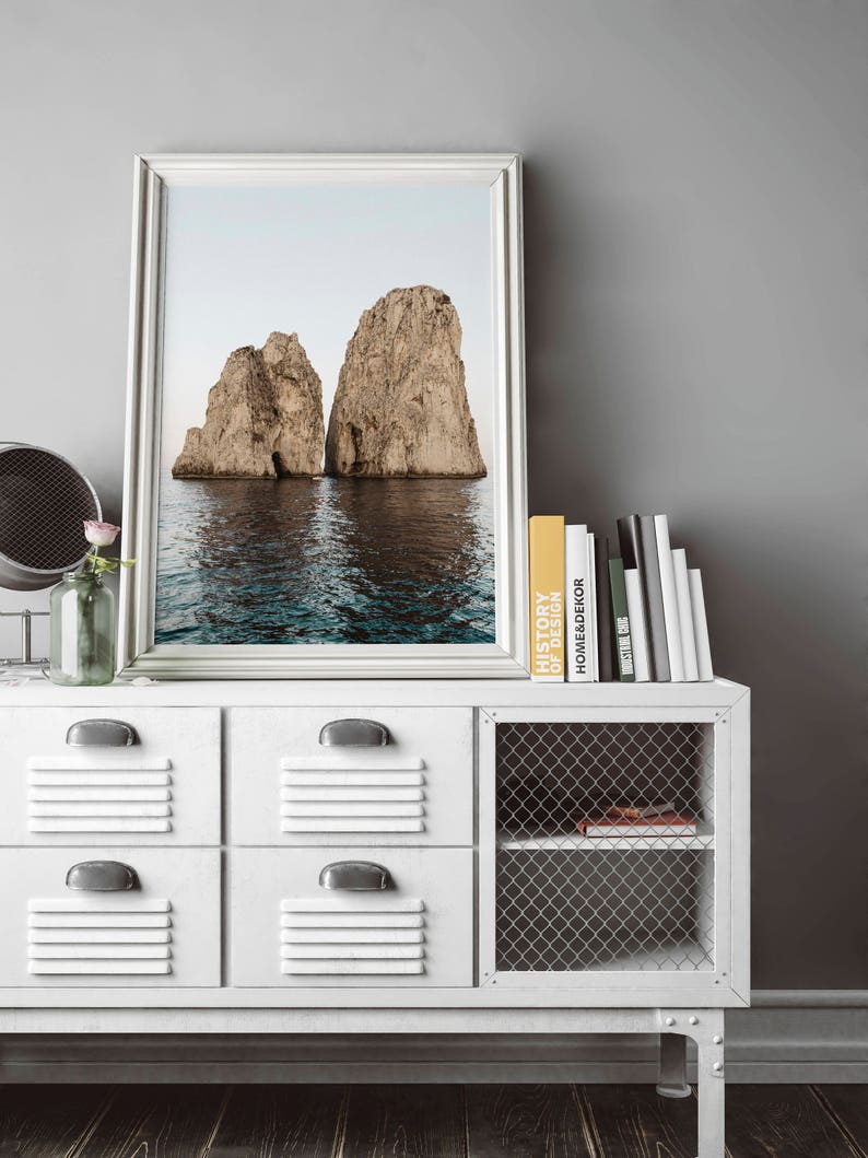 May include: A framed photograph of two large rock formations rising out of the ocean. The water is a deep blue and the sky is a light blue. The photo is hanging on a white wall above a white dresser with a wire mesh shelf.