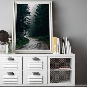 May include: A framed photograph of a winding road through a dense, misty forest. The road is wet, reflecting the overcast sky. The photograph is displayed on a white cabinet with four drawers and a wire mesh door, alongside books and a small mirror.