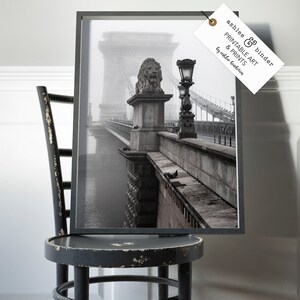 May include: A black and white photograph of a bridge with a lion statue on top. The bridge is covered in fog and the water below is visible. The photo is framed in a black frame.