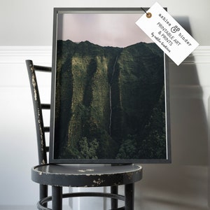 May include: A framed photograph of a lush green mountain range with a waterfall cascading down the side. The photograph is in a black frame and is sitting on a black wooden chair.