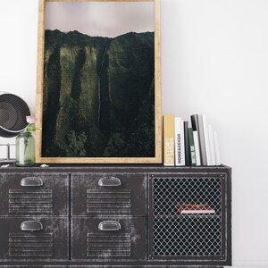 May include: A framed photograph of a lush green mountain range with a waterfall cascading down the side. The photograph is displayed on a dark gray metal cabinet with a wire mesh door. Several books are stacked on top of the cabinet.