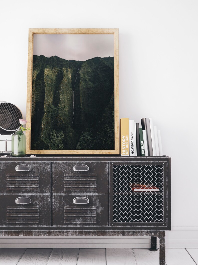 May include: A framed photograph of a lush green mountain range with a waterfall cascading down the side. The photograph is displayed on a dark gray metal cabinet with a wire mesh door. Several books are stacked on top of the cabinet.