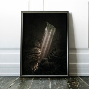 Op de afbeelding: Ingelijste print van een grotinterieur met dramatische lichtstralen. De afbeelding toont een donkere, holle ruimte met zonlicht dat door een opening schijnt en de rotsformaties verlicht. De print wordt weergegeven op een donkere houten vloer tegen een witte muur.
