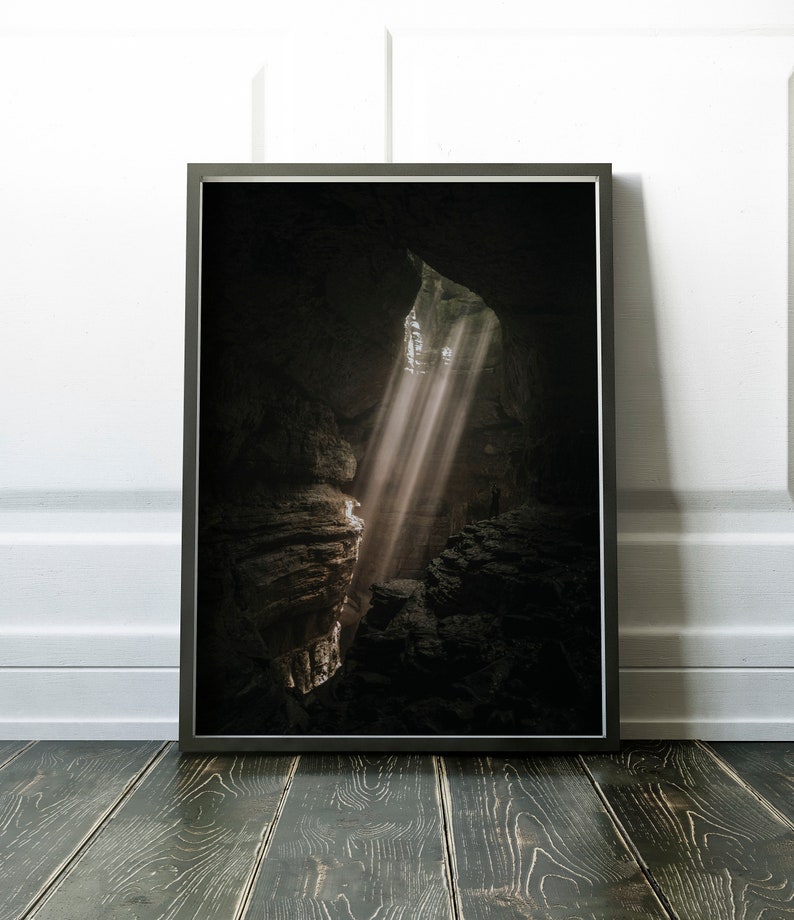 Op de afbeelding: Ingelijste print van een grotinterieur met dramatische lichtstralen. De afbeelding toont een donkere, holle ruimte met zonlicht dat door een opening schijnt en de rotsformaties verlicht. De print wordt weergegeven op een donkere houten vloer tegen een witte muur.