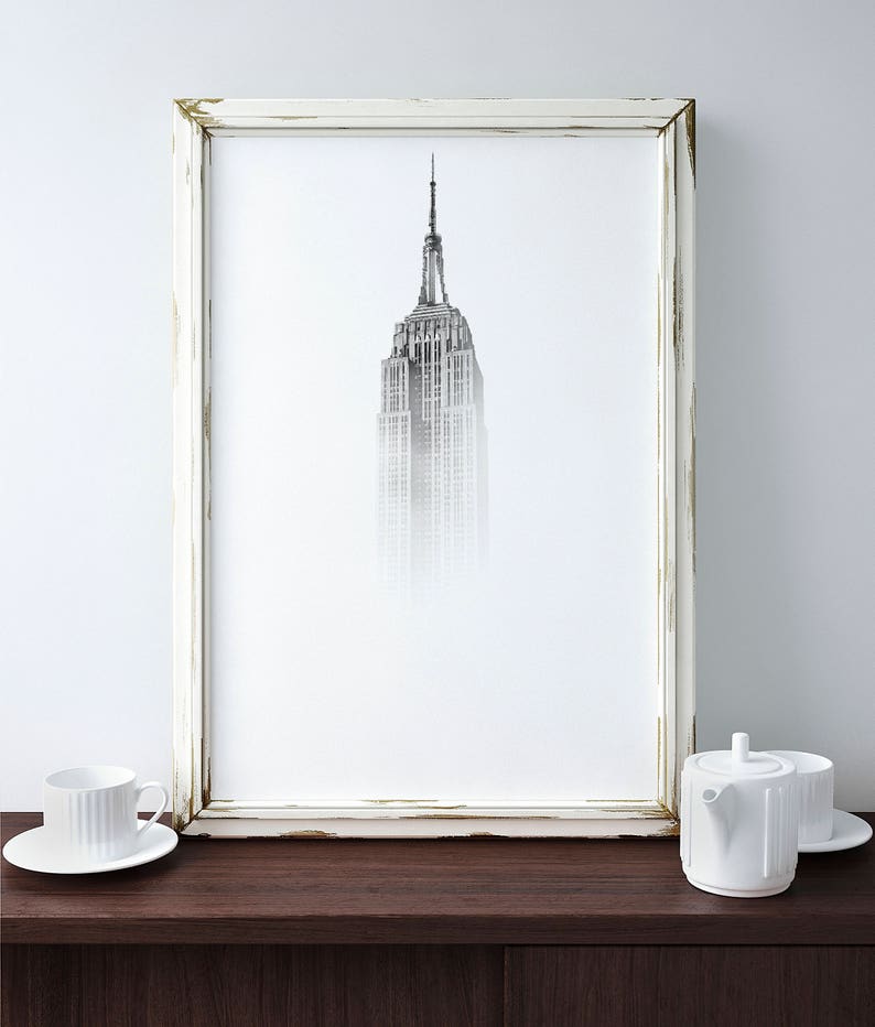 Peut inclure: Une photographie en noir et blanc de l'Empire State Building &agrave; New York, encadr&eacute;e dans un cadre blanc vieilli. Le b&acirc;timent est partiellement obscurci par le brouillard, cr&eacute;ant un effet r&ecirc;veur et &eacute;th&eacute;r&eacute;. Le cadre est pos&eacute; sur une surface en bois brun fonc&eacute;.