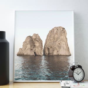 May include: A framed photograph of two large rock formations rising out of the ocean. The water is a light blue and the sky is a pale gray. The rocks are a light brown color.