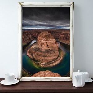 Puede incluir: Fotograf&iacute;a enmarcada del paisaje de Horseshoe Bend, Arizona, con un cielo dram&aacute;tico y el r&iacute;o Colorado. La imagen est&aacute; en un marco blanco envejecido. Una taza y una tetera blancas est&aacute;n sobre una superficie de madera oscura.