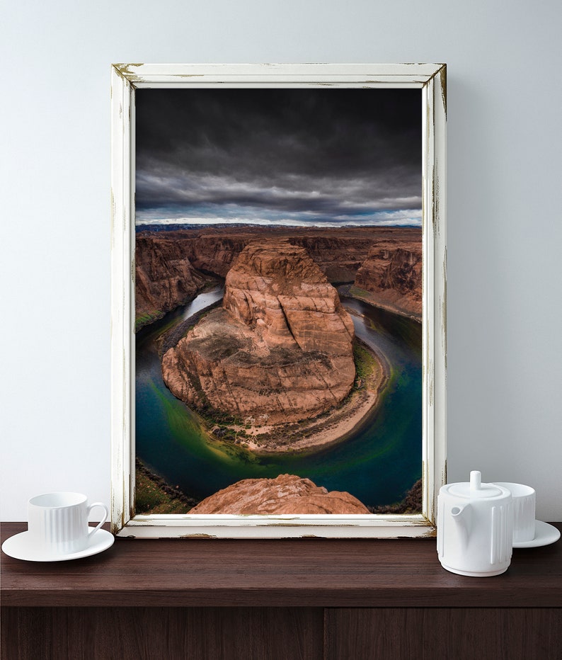 Puede incluir: Fotograf&iacute;a enmarcada del paisaje de Horseshoe Bend, Arizona, con un cielo dram&aacute;tico y el r&iacute;o Colorado. La imagen est&aacute; en un marco blanco envejecido. Una taza y una tetera blancas est&aacute;n sobre una superficie de madera oscura.