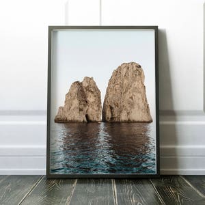 May include: A framed photograph of two large rock formations rising out of the ocean. The rocks are a light brown color and have a rough texture. The water is a deep blue color and is calm.