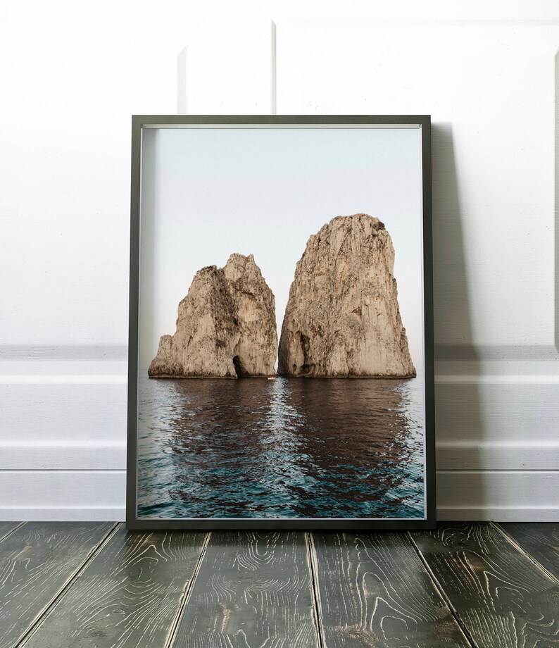 May include: A framed photograph of two large rock formations rising out of the ocean. The rocks are a light brown color and have a rough texture. The water is a deep blue color and is calm.