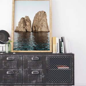 May include: A framed photograph of two large rock formations rising out of the ocean. The photo is hanging on a wall above a black metal cabinet with a wire mesh door.