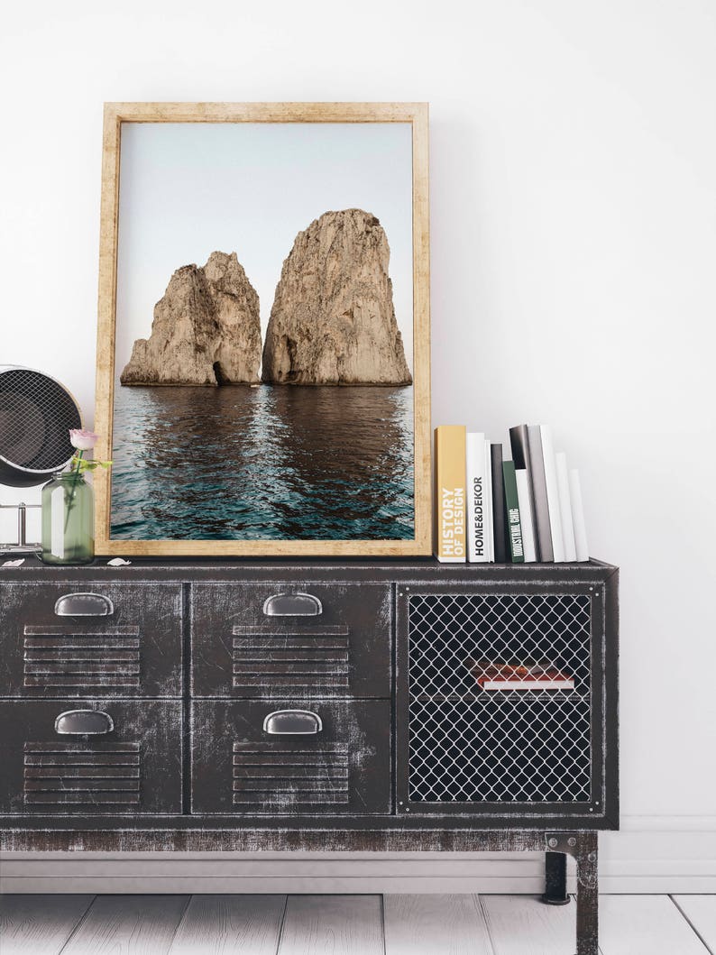 May include: A framed photograph of two large rock formations rising out of the ocean. The photo is hanging on a wall above a black metal cabinet with a wire mesh door.