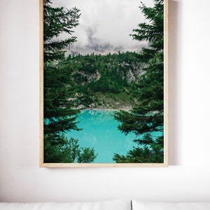 Op de afbeelding: Een ingelijste foto van een bergmeer omringd door bomen. Het meer is een levendige turquoise blauw en de bergen zijn bedekt met mist. De bomen zijn groen en omlijsten het meer.