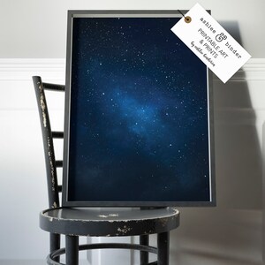K&ouml;nnte beinhalten: Ein gerahmter Druck eines dunkelblauen Nachthimmels mit einer Galaxie aus wei&szlig;en Sternen. Der Druck h&auml;ngt an einem schwarzen Stuhl mit einer wei&szlig;en Wand im Hintergrund. Der Druck hat ein Etikett, das an ihm befestigt ist und "ashlee & binder PRINTABLE ART & PRINTS by ashlee binder" lautet.