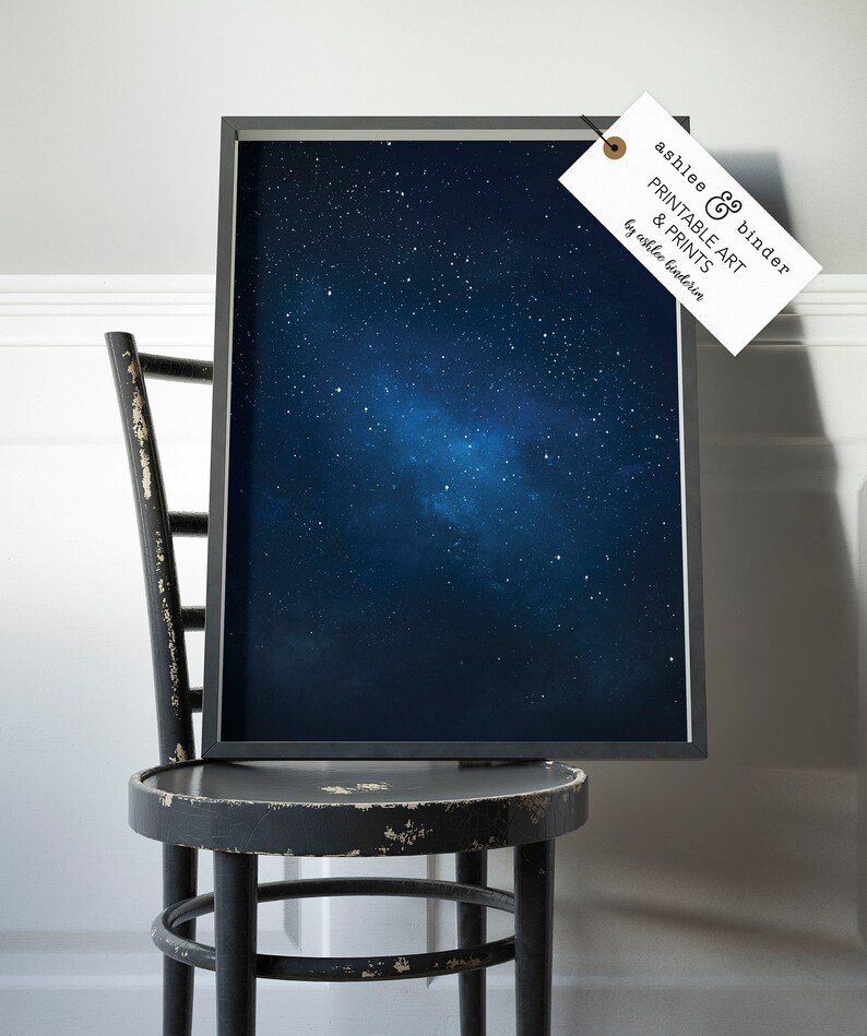 K&ouml;nnte beinhalten: Ein gerahmter Druck eines dunkelblauen Nachthimmels mit einer Galaxie aus wei&szlig;en Sternen. Der Druck h&auml;ngt an einem schwarzen Stuhl mit einer wei&szlig;en Wand im Hintergrund. Der Druck hat ein Etikett, das an ihm befestigt ist und "ashlee & binder PRINTABLE ART & PRINTS by ashlee binder" lautet.