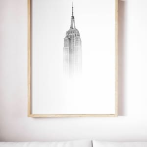 Peut inclure: Une photographie en noir et blanc de l'Empire State Building &agrave; New York. Le b&acirc;timent est encadr&eacute; dans un cadre en bois clair et est accroch&eacute; &agrave; un mur blanc.