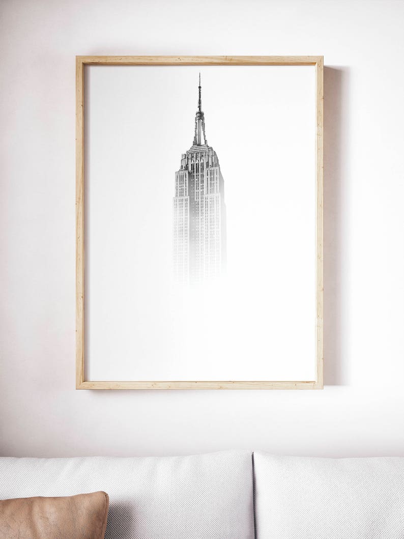 Peut inclure: Une photographie en noir et blanc de l'Empire State Building &agrave; New York. Le b&acirc;timent est encadr&eacute; dans un cadre en bois clair et est accroch&eacute; &agrave; un mur blanc.