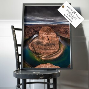 Puede incluir: Impresi&oacute;n enmarcada de Horseshoe Bend, Arizona, con un paisaje dram&aacute;tico con un r&iacute;o que serpentea alrededor de una formaci&oacute;n de arenisca. La impresi&oacute;n est&aacute; en un marco negro y se muestra en una silla negra desgastada. La etiqueta dice "ashlee & binder PRINTABLE ART & PRINTS by ashlee binderia."