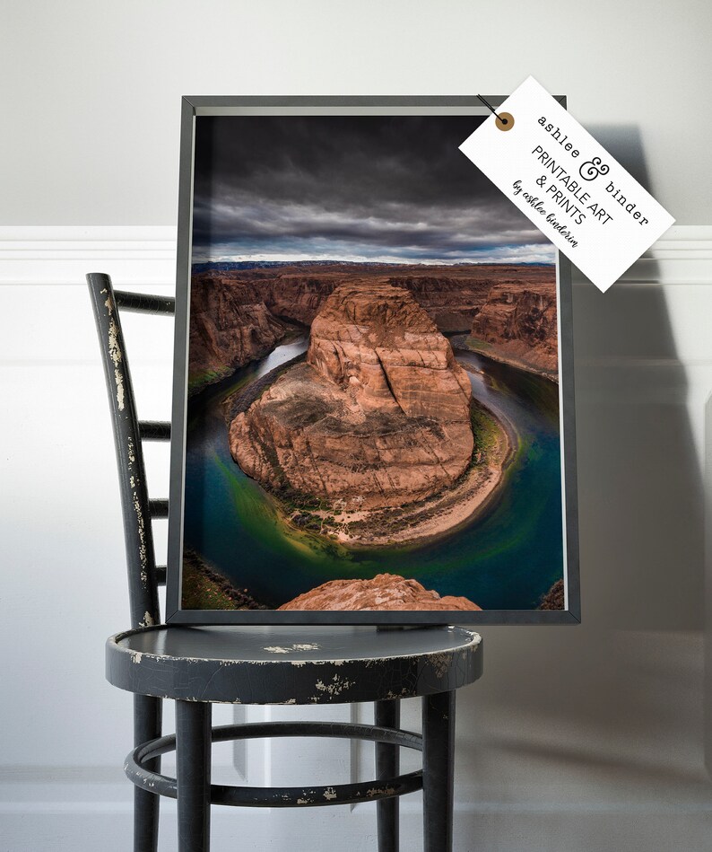 Puede incluir: Impresi&oacute;n enmarcada de Horseshoe Bend, Arizona, con un paisaje dram&aacute;tico con un r&iacute;o que serpentea alrededor de una formaci&oacute;n de arenisca. La impresi&oacute;n est&aacute; en un marco negro y se muestra en una silla negra desgastada. La etiqueta dice "ashlee & binder PRINTABLE ART & PRINTS by ashlee binderia."