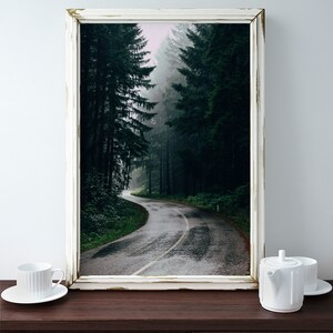 May include: A framed print of a winding road through a dense forest, with tall, dark green trees lining the path. The road is wet, and the sky is overcast. The frame is white with a distressed finish. A white teacup and teapot are on a dark wooden surface.
