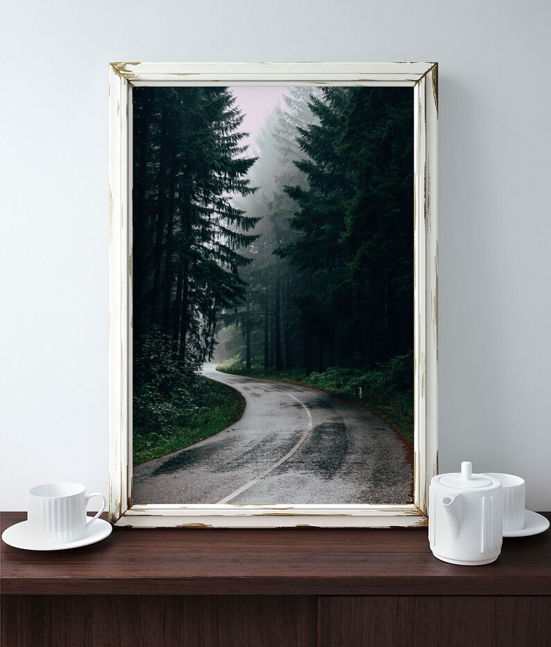 May include: A framed print of a winding road through a dense forest, with tall, dark green trees lining the path. The road is wet, and the sky is overcast. The frame is white with a distressed finish. A white teacup and teapot are on a dark wooden surface.