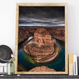 Puede incluir: Impresi&oacute;n enmarcada de Horseshoe Bend, Arizona, que muestra la dram&aacute;tica curva en forma de herradura del r&iacute;o Colorado. La imagen presenta un r&iacute;o azul-verdoso vibrante contra formaciones rocosas rojas bajo un cielo dram&aacute;tico y nublado. El marco es de color madera clara.