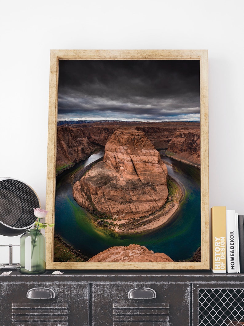 Puede incluir: Impresi&oacute;n enmarcada de Horseshoe Bend, Arizona, que muestra la dram&aacute;tica curva en forma de herradura del r&iacute;o Colorado. La imagen presenta un r&iacute;o azul-verdoso vibrante contra formaciones rocosas rojas bajo un cielo dram&aacute;tico y nublado. El marco es de color madera clara.