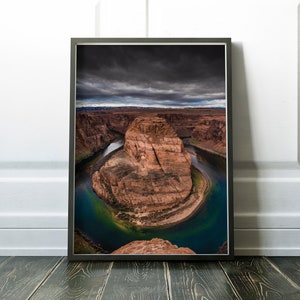 Puede incluir: Fotograf&iacute;a enmarcada del paisaje de Horseshoe Bend, Arizona. La imagen captura la ic&oacute;nica curva del r&iacute;o Colorado, mostrando las formaciones rocosas rojas y las aguas turquesas bajo un cielo dram&aacute;tico y nublado. La impresi&oacute;n enmarcada se exhibe sobre un suelo de madera.