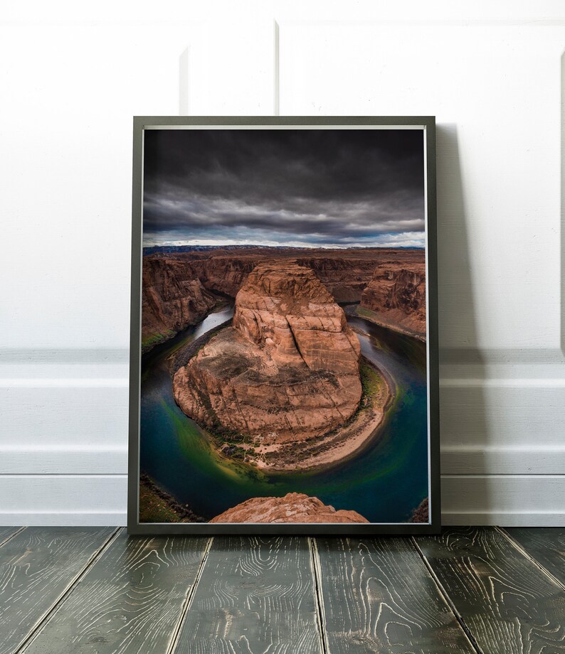 Puede incluir: Fotograf&iacute;a enmarcada del paisaje de Horseshoe Bend, Arizona. La imagen captura la ic&oacute;nica curva del r&iacute;o Colorado, mostrando las formaciones rocosas rojas y las aguas turquesas bajo un cielo dram&aacute;tico y nublado. La impresi&oacute;n enmarcada se exhibe sobre un suelo de madera.
