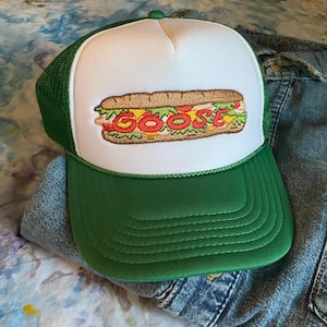 May include: A green and white trucker hat featuring a sandwich design with the word "GOOSE" spelled out in red, yellow, and green. The hat has a green brim and mesh back, and a white front panel.